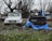 Two Suzuki vehicles are positioned side by side on a grassy area, with one being a white SUV and the other a blue convertible. The blue convertible is partially covered with a tattered blue tarp. The ground is scattered with fallen leaves, and barren trees without foliage are visible in the background.