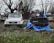 Two Suzuki vehicles are positioned side by side on a grassy area, with one being a white SUV and the other a blue convertible. The blue convertible is partially covered with a tattered blue tarp. The ground is scattered with fallen leaves, and barren trees without foliage are visible in the background.