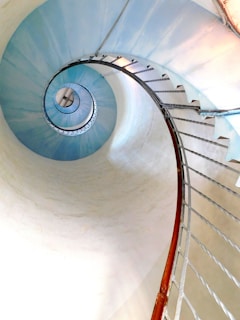 A spiraling staircase with a metal railing curves upwards in a smooth, continuous motion, creating a mesmerizing spiral effect. The stairs are painted in a soft blue color that contrasts with the white background, and the lighting adds a gentle gradient, enhancing the depth of the spiral.