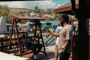 A person holding a camera on a tripod is standing on a wooden deck overlooking a scenic area with hills and colorful buildings. Another person is sitting relaxed on a wooden swing. The setting appears to be sunny and leisurely.
