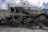 A collapsed and partially destroyed building with exposed structural beams and rubble scattered in front. The facade shows significant damage, with some parts still hanging precariously. Vegetation is visible among the debris.