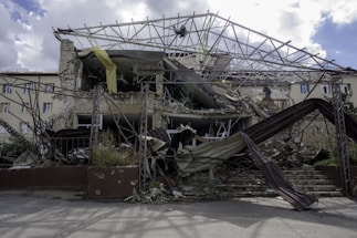 A collapsed and partially destroyed building with exposed structural beams and rubble scattered in front. The facade shows significant damage, with some parts still hanging precariously. Vegetation is visible among the debris.