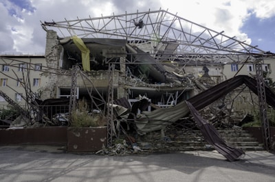 A collapsed and partially destroyed building with exposed structural beams and rubble scattered in front. The facade shows significant damage, with some parts still hanging precariously. Vegetation is visible among the debris.
