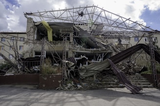 A collapsed and partially destroyed building with exposed structural beams and rubble scattered in front. The facade shows significant damage, with some parts still hanging precariously. Vegetation is visible among the debris.