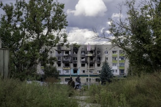 A damaged multi-story residential building stands in the background, with visible burn marks and broken windows. The building is surrounded by green trees and overgrown grass. Two people are visible in the distance, moving toward the building. Overhead, the sky is filled with dark, stormy clouds.