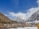 A village nested in a valley with snow-capped mountains in the background. The foreground features colorful prayer flags and traditional stone houses, while the clear blue sky enhances the scenic beauty.