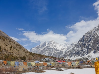 A village nested in a valley with snow-capped mountains in the background. The foreground features colorful prayer flags and traditional stone houses, while the clear blue sky enhances the scenic beauty.