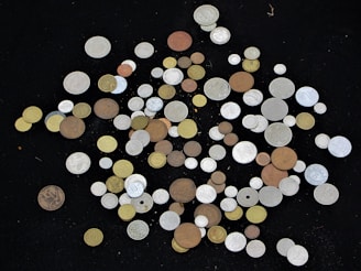 A scattered collection of coins of various sizes, shapes, and colors displayed against a black background. The coins include copper, silver, and gold hues, with some ornate designs and diverse inscriptions visible.
