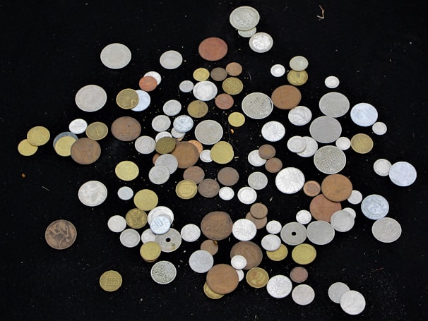 A scattered collection of coins of various sizes, shapes, and colors displayed against a black background. The coins include copper, silver, and gold hues, with some ornate designs and diverse inscriptions visible.
