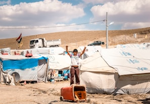 A young boy stands with raised arms in front of a row of tents in a refugee camp. The tents have UNHCR logos, indicating the involvement of the United Nations High Commissioner for Refugees. In the background, a truck and a few vehicles are visible alongside a hillside. A national flag is flying on a pole, and the ground is dry and dusty.
