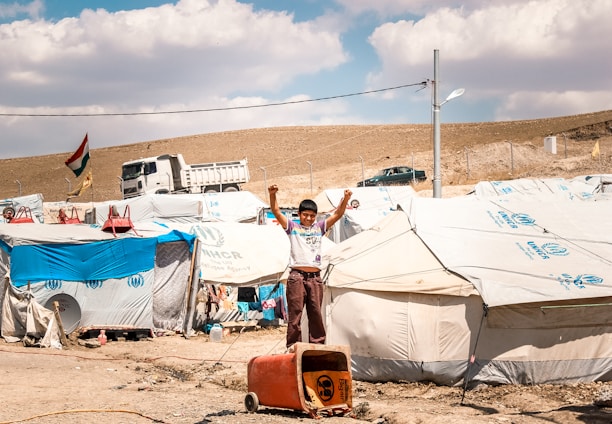 A young boy stands with raised arms in front of a row of tents in a refugee camp. The tents have UNHCR logos, indicating the involvement of the United Nations High Commissioner for Refugees. In the background, a truck and a few vehicles are visible alongside a hillside. A national flag is flying on a pole, and the ground is dry and dusty.