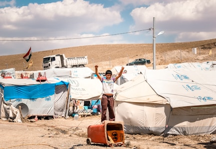 A young boy stands with raised arms in front of a row of tents in a refugee camp. The tents have UNHCR logos, indicating the involvement of the United Nations High Commissioner for Refugees. In the background, a truck and a few vehicles are visible alongside a hillside. A national flag is flying on a pole, and the ground is dry and dusty.