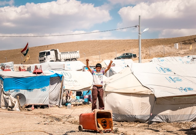 A young boy stands with raised arms in front of a row of tents in a refugee camp. The tents have UNHCR logos, indicating the involvement of the United Nations High Commissioner for Refugees. In the background, a truck and a few vehicles are visible alongside a hillside. A national flag is flying on a pole, and the ground is dry and dusty.
