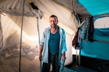 A man stands inside a tent with an expression of weariness and determination. The tent is modestly furnished with pillows and clothes hanging on a makeshift line. The lighting suggests it is daytime, and the overall setting appears to be temporary and basic.