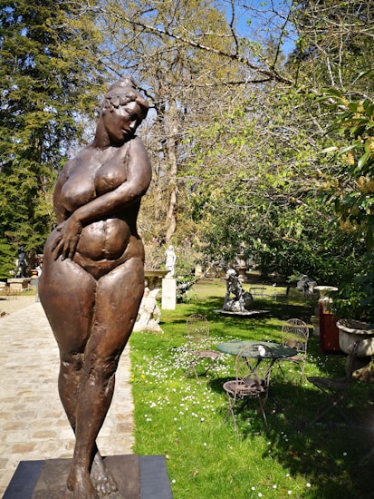 A bronze sculpture of a woman stands in a garden setting. The sculpture depicts a full-figured, nude female in a relaxed pose. Surrounding the sculpture are green lawns, trees with sparse leaves, and garden furniture. The scene conveys a sense of tranquility and natural beauty.