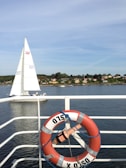 A scenic view showing a sailing boat on a calm body of water, with a shoreline featuring small houses and trees in the background. In the foreground, a red and white lifebuoy with the word 'OSLO' is attached to a railing.