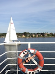 A scenic view showing a sailing boat on a calm body of water, with a shoreline featuring small houses and trees in the background. In the foreground, a red and white lifebuoy with the word 'OSLO' is attached to a railing.