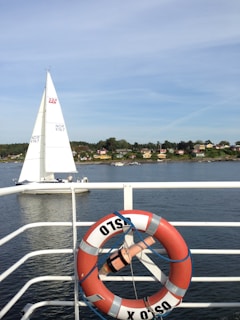 A scenic view showing a sailing boat on a calm body of water, with a shoreline featuring small houses and trees in the background. In the foreground, a red and white lifebuoy with the word 'OSLO' is attached to a railing.