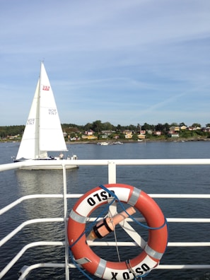 A scenic view showing a sailing boat on a calm body of water, with a shoreline featuring small houses and trees in the background. In the foreground, a red and white lifebuoy with the word 'OSLO' is attached to a railing.