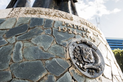 A stone monument with engraved text and an ornate metal emblem. The text includes the years 1924-1960, and the emblem features the Dominican Republic seal. Sunlight casts shadows, adding depth to the textured surface.