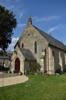 A quaint stone church with a pitched roof set against a clear blue sky, surrounded by a well-manicured lawn dotted with white flowers. The building features arched doorways and windows, accented with dark wooden doors and slate roofing. Flanking the entrance are two potted plants, and a pathway leads up to the entrance, creating an inviting atmosphere.