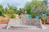 A stone monument featuring a bust of a man is situated in a park setting. The base of the monument is constructed with patterned stone tiles, arranged in a semicircular platform. Surrounding the monument are lush green trees and a bright turquoise and orange building in the background.