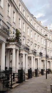 A row of elegant terraced houses with white columns and black wrought iron railings. Each house features multiple levels, balconies, and large windows. A single street lamp stands along the paved walkway in front of the houses.