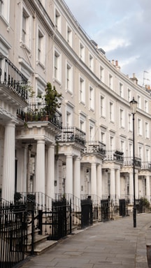 A row of elegant terraced houses with white columns and black wrought iron railings. Each house features multiple levels, balconies, and large windows. A single street lamp stands along the paved walkway in front of the houses.