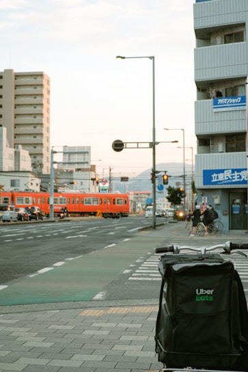 An urban scene featuring a street with a bicycle and an Uber Eats delivery bag in the foreground. A red train is visible in the background, crossing a level crossing on the left. The surroundings include several tall buildings, street lamps, and cyclists. The sky appears cloudy.