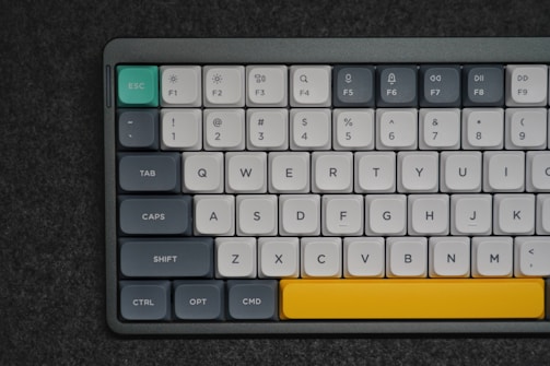 A mechanical keyboard with various colored keys, predominantly white with gray, teal, and a standout yellow spacebar. The keys include function keys labeled F1 to F9, as well as standard keyboard letters and symbols. The keyboard is set against a dark, textured surface.