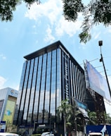A modern high-rise building with a glass facade, reflecting the bright blue sky and scattered white clouds. The building has prominent signage for a coworking space, located in an urban area with visible trees and a large billboard. Vehicles are parked nearby, indicating an accessible location.
