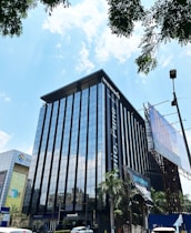 A modern high-rise building with a glass facade, reflecting the bright blue sky and scattered white clouds. The building has prominent signage for a coworking space, located in an urban area with visible trees and a large billboard. Vehicles are parked nearby, indicating an accessible location.