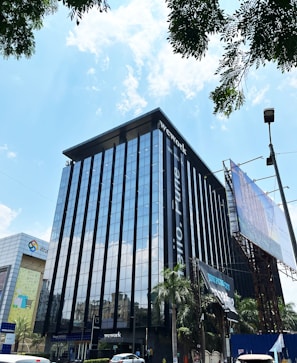 A modern high-rise building with a glass facade, reflecting the bright blue sky and scattered white clouds. The building has prominent signage for a coworking space, located in an urban area with visible trees and a large billboard. Vehicles are parked nearby, indicating an accessible location.