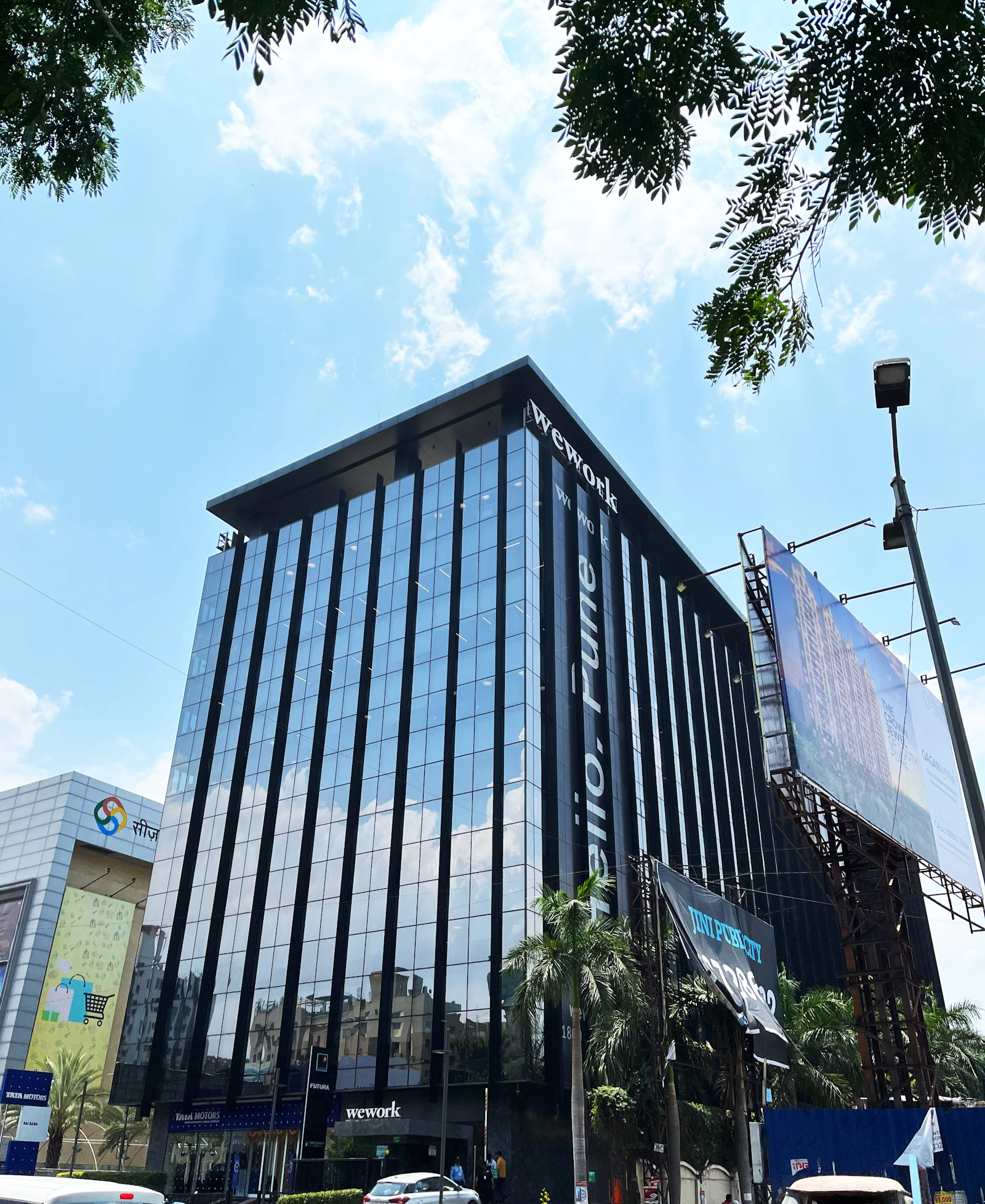A modern high-rise building with a glass facade, reflecting the bright blue sky and scattered white clouds. The building has prominent signage for a coworking space, located in an urban area with visible trees and a large billboard. Vehicles are parked nearby, indicating an accessible location.
