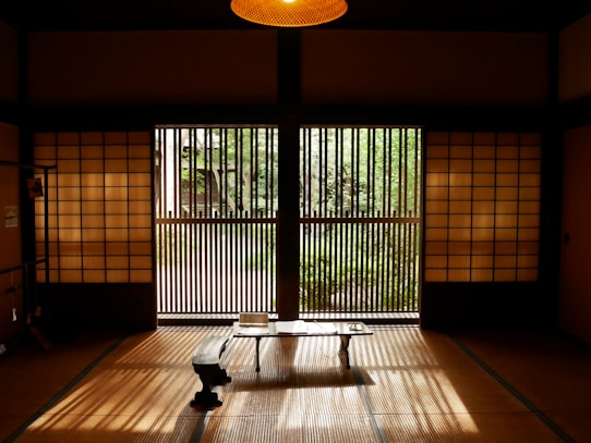 A traditional Japanese room featuring shoji screens and tatami flooring. Natural light filters through wooden slatted doors overlooking a garden with greenery visible outside. The interior includes a minimalist setting with a low table and a small bench, creating an atmosphere of calm and simplicity.