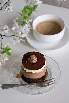 A layered chocolate dessert with a small chocolate ball on top sits on a decorative glass plate. Next to the dessert, there is a fork on the plate. In the background is a white cup of coffee with creamy foam on a white saucer. A branch of white flowers with green leaves adds a touch of nature to the scene.