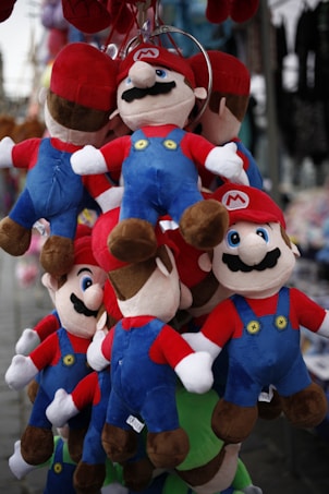 Several plush dolls wearing red hats, blue overalls, and red shirts are hanging from a display rack. They have black mustaches, white gloves, and brown shoes. The dolls are grouped closely together, creating a colorful and playful assortment.