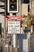 A traffic light mounted on a pole is accompanied by a sign in both Arabic and English that reads 'Keep Crossing Clear.' The setting appears to be an urban street with palm trees and modern buildings in the background.