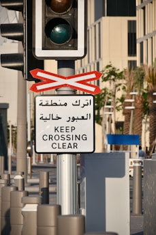 A traffic light mounted on a pole is accompanied by a sign in both Arabic and English that reads 'Keep Crossing Clear.' The setting appears to be an urban street with palm trees and modern buildings in the background.