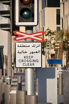A traffic light mounted on a pole is accompanied by a sign in both Arabic and English that reads 'Keep Crossing Clear.' The setting appears to be an urban street with palm trees and modern buildings in the background.