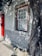 A quaint exterior of a building with a red door and window frames. The facade is decorated with patterned tiles, featuring floral motifs. Sunlight trickles through the leaves of a large tree, casting intricate shadows on the wall and cobblestone path.