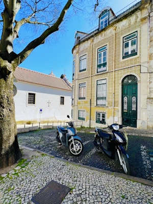 Two motorbikes are parked on a cobblestone street in front of a historic building with intricate tile work. The building has several windows and a large arched door. A tree with bare branches extends over the scene, and a white building with a tiled roof is visible in the background under a clear blue sky.