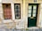 A rustic facade featuring a dark green door with four glass panels and a golden mail slot. Adjacent to the door are two windows with black and brown metal bars against a stone and plaster wall. A blue street number sign with the number 2 is above the door. The ground is cobblestone with some patches of greenery.
