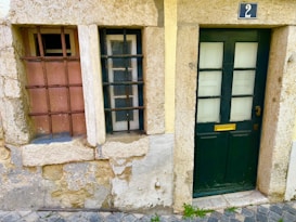 A rustic facade featuring a dark green door with four glass panels and a golden mail slot. Adjacent to the door are two windows with black and brown metal bars against a stone and plaster wall. A blue street number sign with the number 2 is above the door. The ground is cobblestone with some patches of greenery.