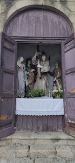 A set of religious statues arranged within a niche on a purple wooden backdrop. Four statues, including a central figure carrying a cross, are surrounded by figures in historical attire. The arrangement is framed by open wooden doors, with a white cloth at the base, alongside plants and a bottle.