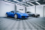 Two luxury sports cars are parked in an elegant, modern showroom with a checkered floor and overhead lighting. The car in the foreground is a vibrant blue with sleek curves, while the car in the background has a metallic silver finish.