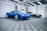 Two luxury sports cars are parked in an elegant, modern showroom with a checkered floor and overhead lighting. The car in the foreground is a vibrant blue with sleek curves, while the car in the background has a metallic silver finish.