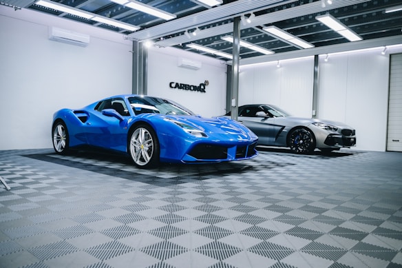 Two luxury sports cars are parked in an elegant, modern showroom with a checkered floor and overhead lighting. The car in the foreground is a vibrant blue with sleek curves, while the car in the background has a metallic silver finish.