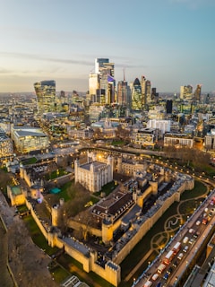 An aerial view of a cityscape featuring a blend of modern skyscrapers and historic architecture. The foreground highlights a medieval castle complex surrounded by walls and green lawns. Bright lights illuminate the area, and a line of vehicles can be seen on the adjacent road.