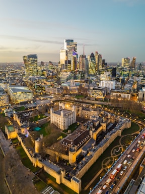 An aerial view of a cityscape featuring a blend of modern skyscrapers and historic architecture. The foreground highlights a medieval castle complex surrounded by walls and green lawns. Bright lights illuminate the area, and a line of vehicles can be seen on the adjacent road.